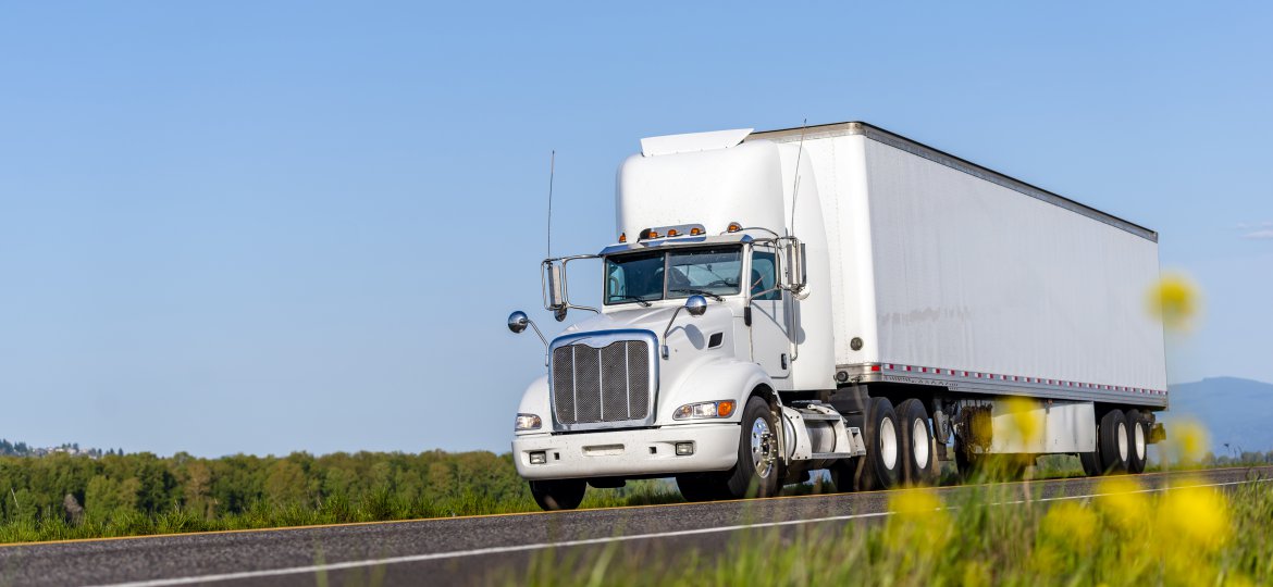 Big rig white day cab semi truck for local deliveries with long box semi trailer driving on the summer road with yellow flowers